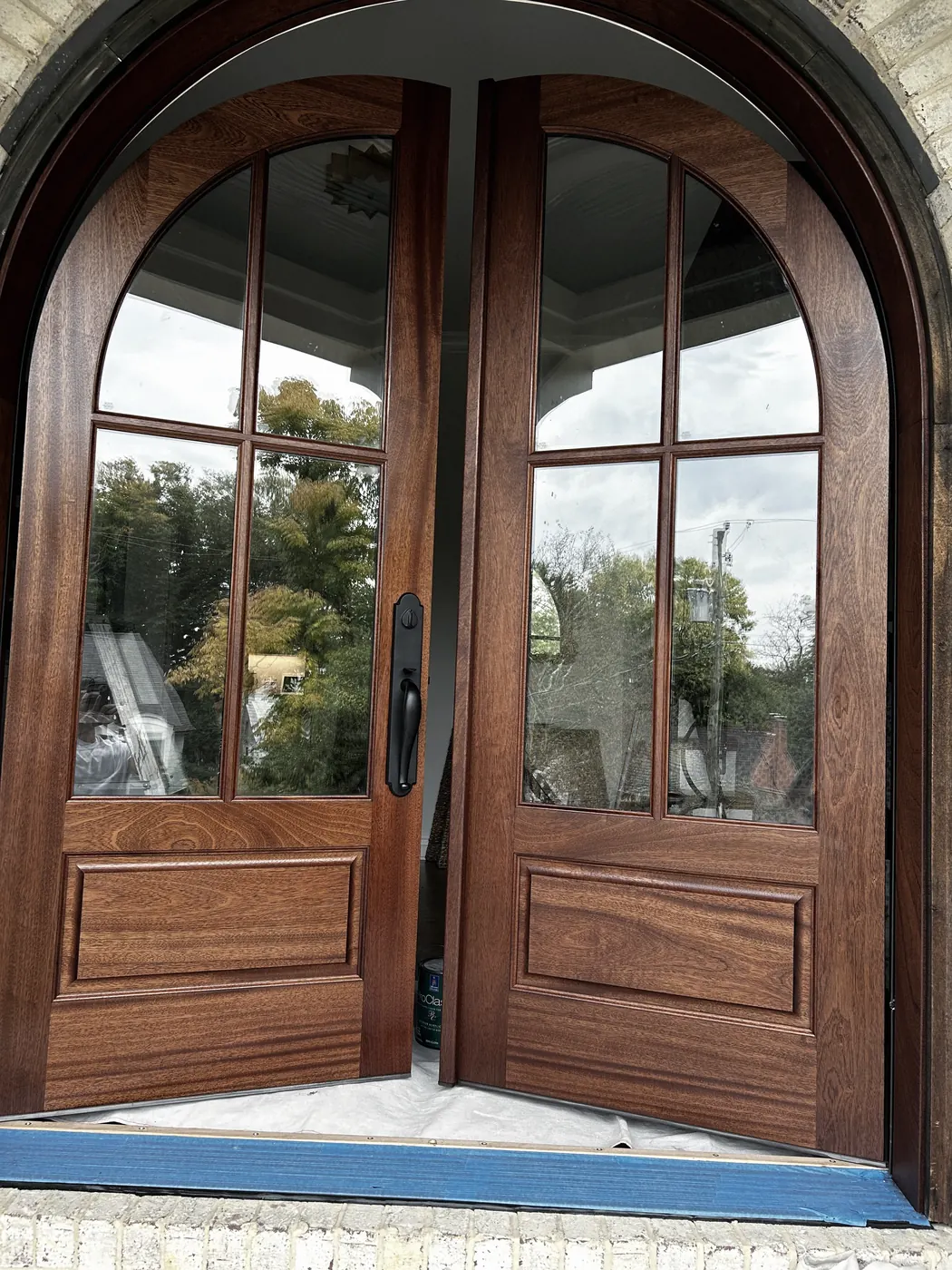 The same arched wood doors with the wood a deep red-brown, standing slightly apart, blue tape and a drop cloth at the sill, trees reflected in the glass.