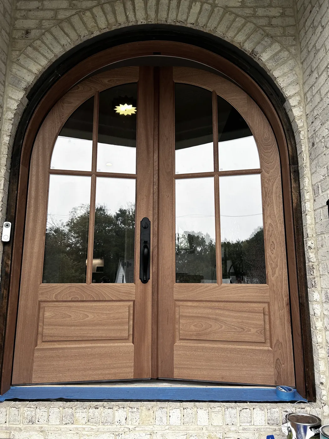Arched pair of wood double doors with glass panes and black handles in a pale brick archway, blue tape along the sill, the wood a light tan.