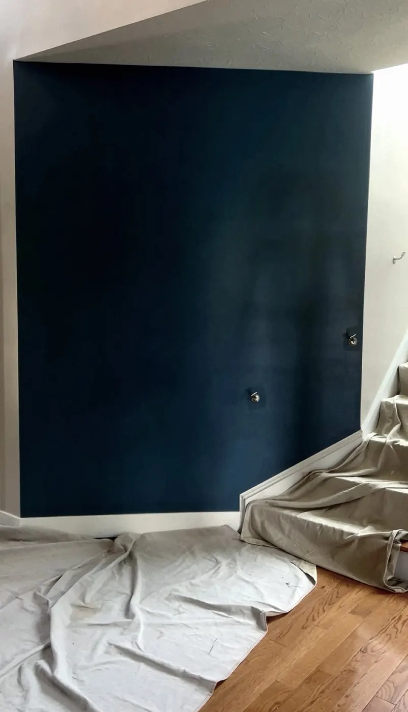 Dark blue accent wall beside a white wall, drop cloths over the hardwood floor and stairs, two wall hooks.