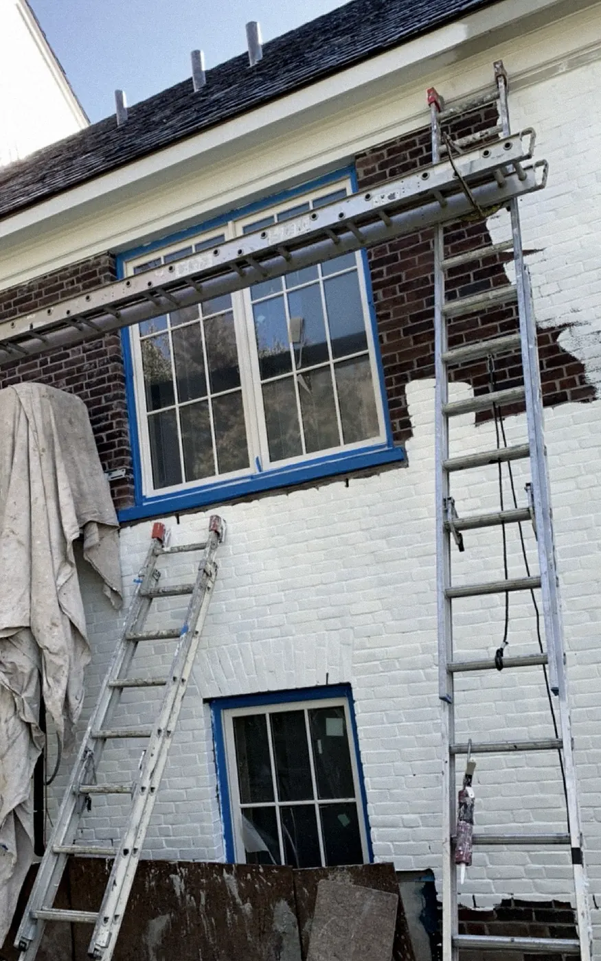 Brick wall painted white from the bottom up, dark red-brown brick still showing around a blue-taped window, two extension ladders and a hanging drop cloth.