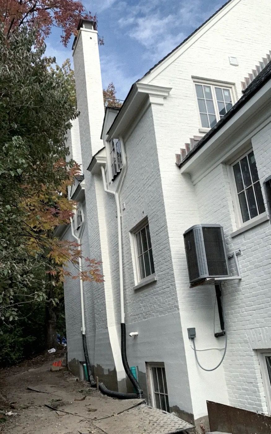Tall side wall of a white-painted brick house with a long chimney, cream window trim and an air conditioner condenser, autumn leaves at the left.