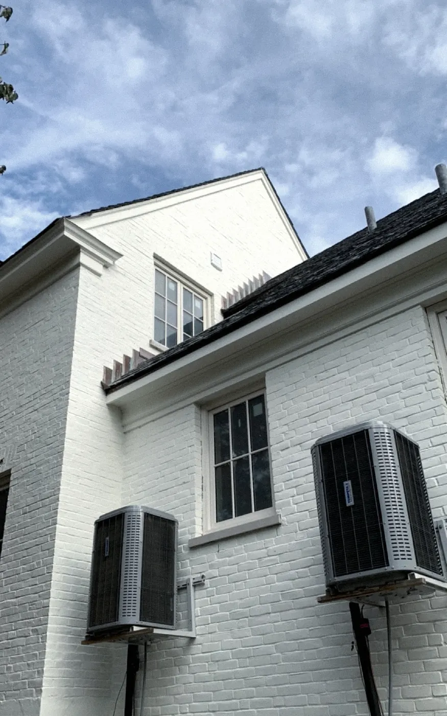 White painted brick wall with cream window trim, two air conditioner condensers on brackets and a gabled roofline against a blue sky.
