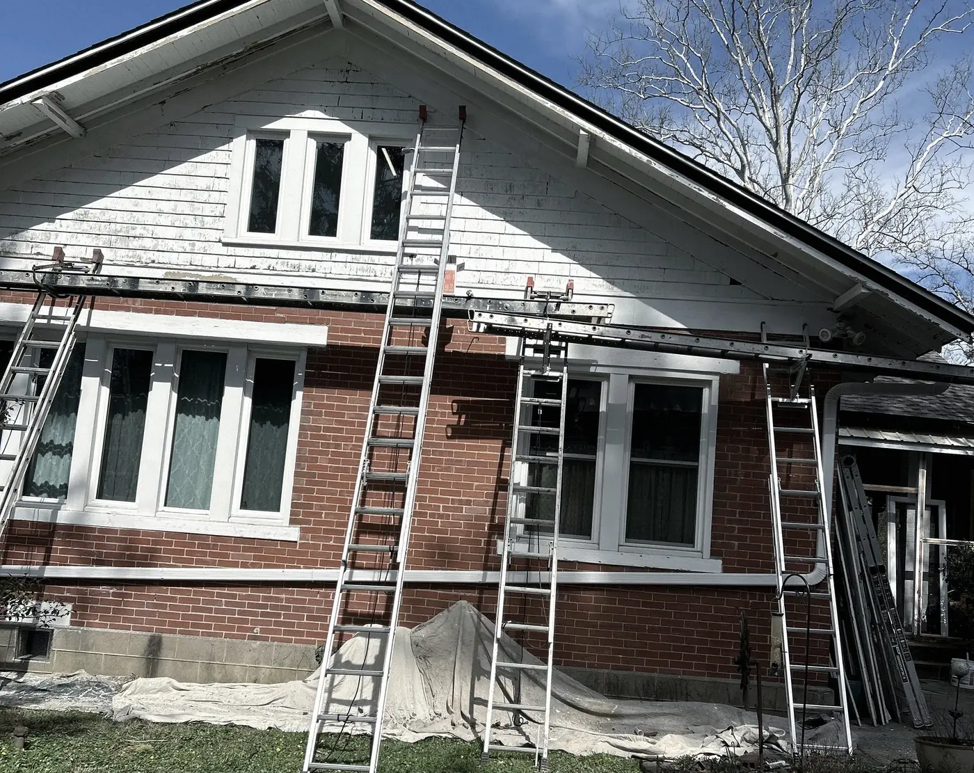 Extension ladders against a brick bungalow, drop cloths along the foundation, the gable scraped.