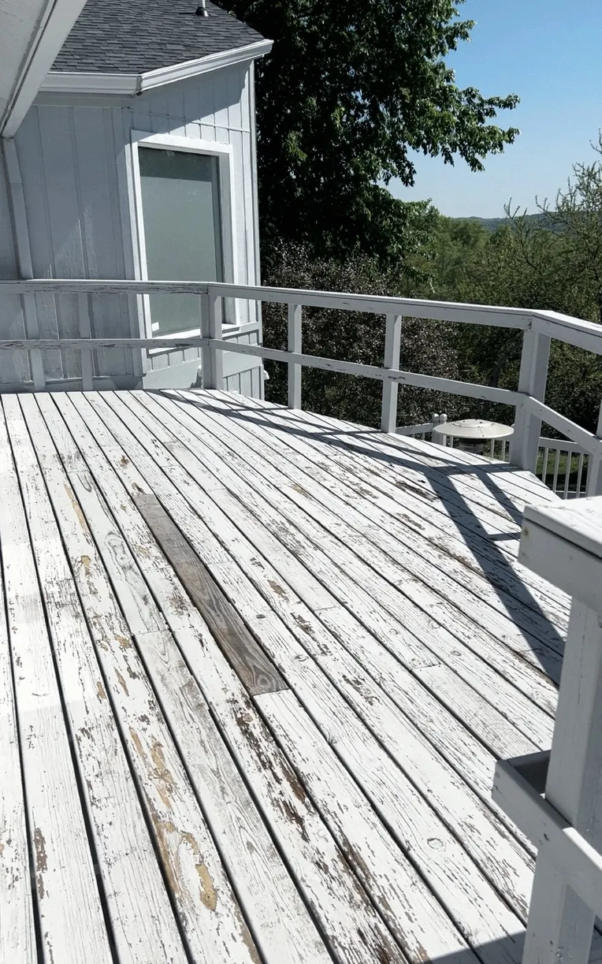 White-painted deck boards peeling and worn to bare wood, one replaced board, a railing and a gray-sided wall.