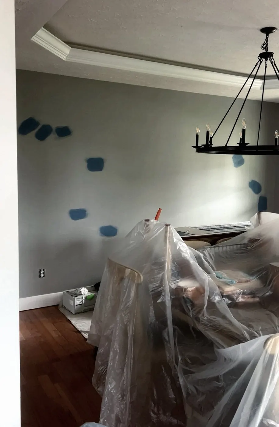 Dining room with furniture wrapped in plastic, an iron chandelier overhead, blue paint swatches on the wall.