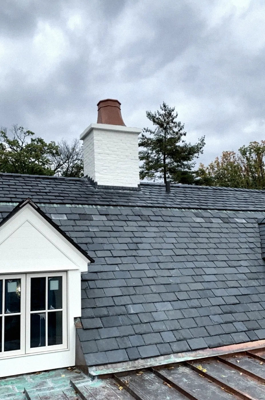 White-painted brick chimney with a copper cap above a gray slate roof, a dormer with a French window at the left, copper roof at the bottom.