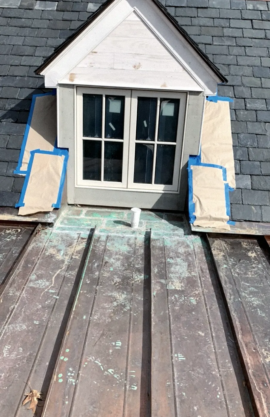 Dormer with a white peaked top and a double window, brown paper and blue tape along both sides, a slate roof behind and a copper roof below.