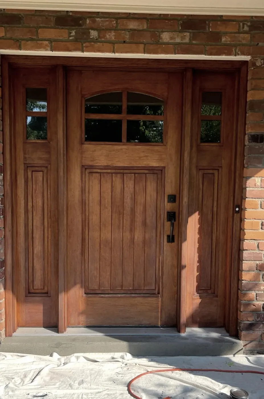 Honey-stained front door with two sidelights in a brick wall, a paint can and orange extension cord on a drop cloth in front.