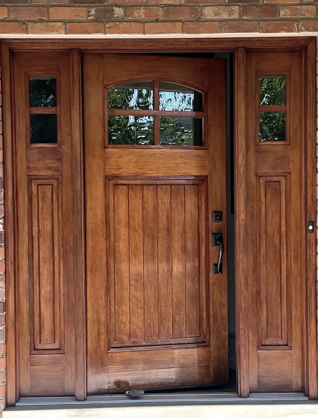 Honey-brown stained front door with two sidelights and an arched glass panel, set in a brick wall, standing slightly open.
