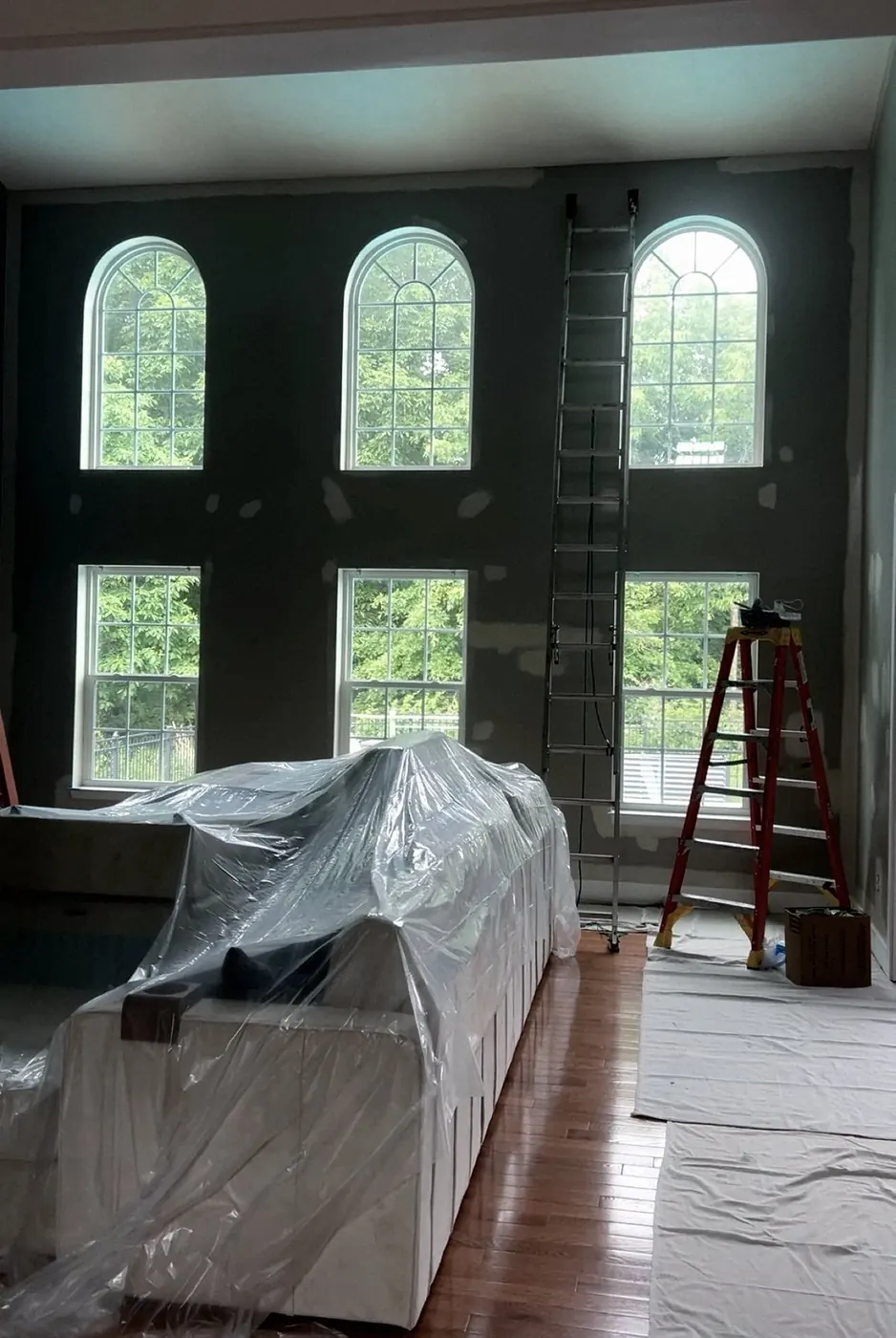 Great room mid-job: the sofa wrapped in plastic, a stepladder at the right, floors covered, arched windows behind.