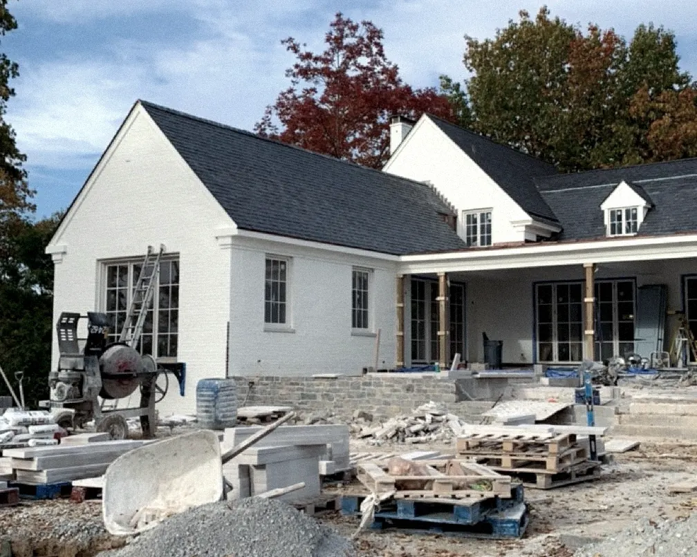 Back of a white-painted brick house with a gray slate roof, dormers and a covered porch on wood posts, building materials and bare dirt in front, autumn trees behind.