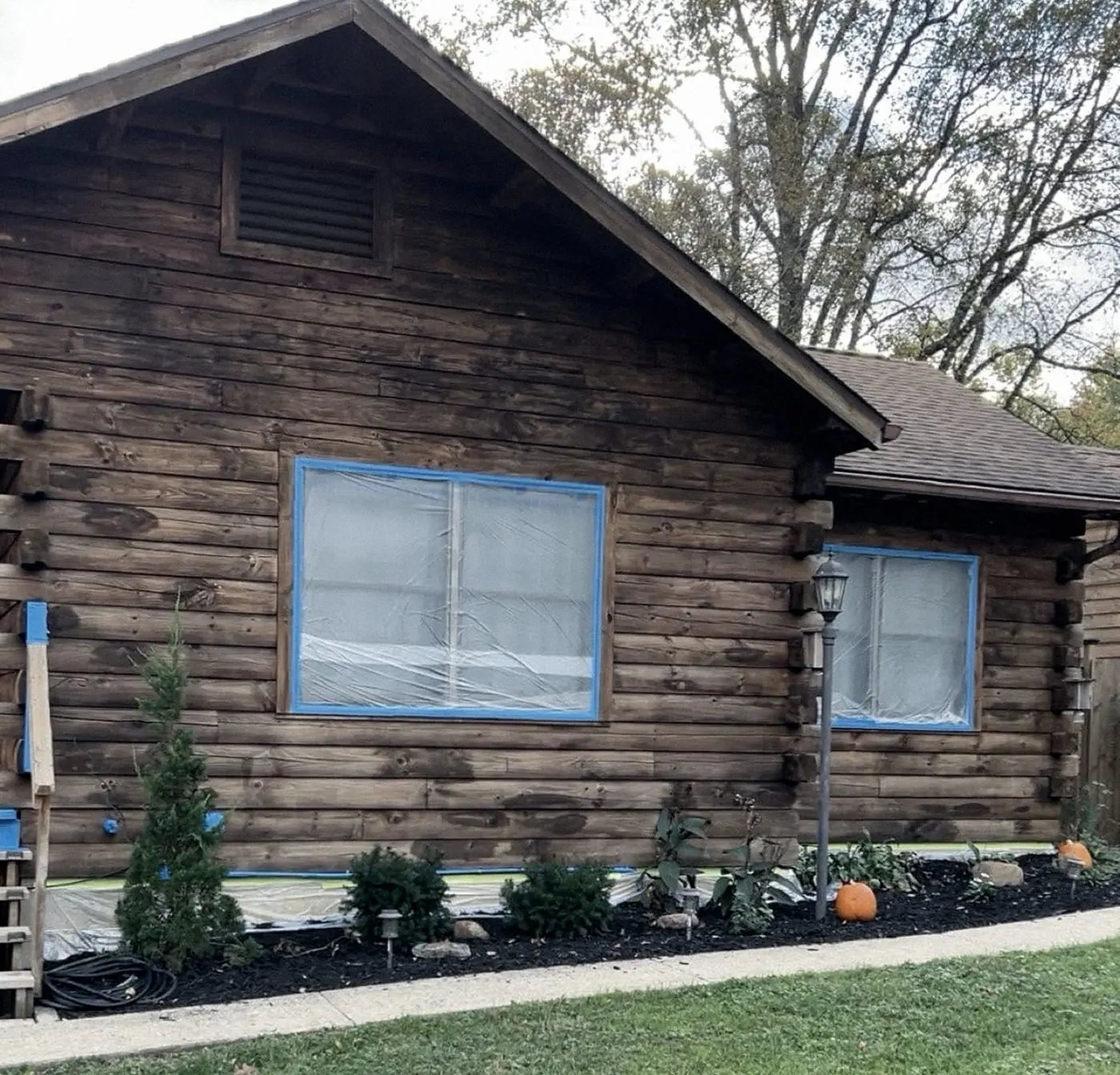 Dark brown log-sided home with a louvered gable vent, windows masked in plastic with blue tape, a lamp post, plantings and pumpkins along the walk.