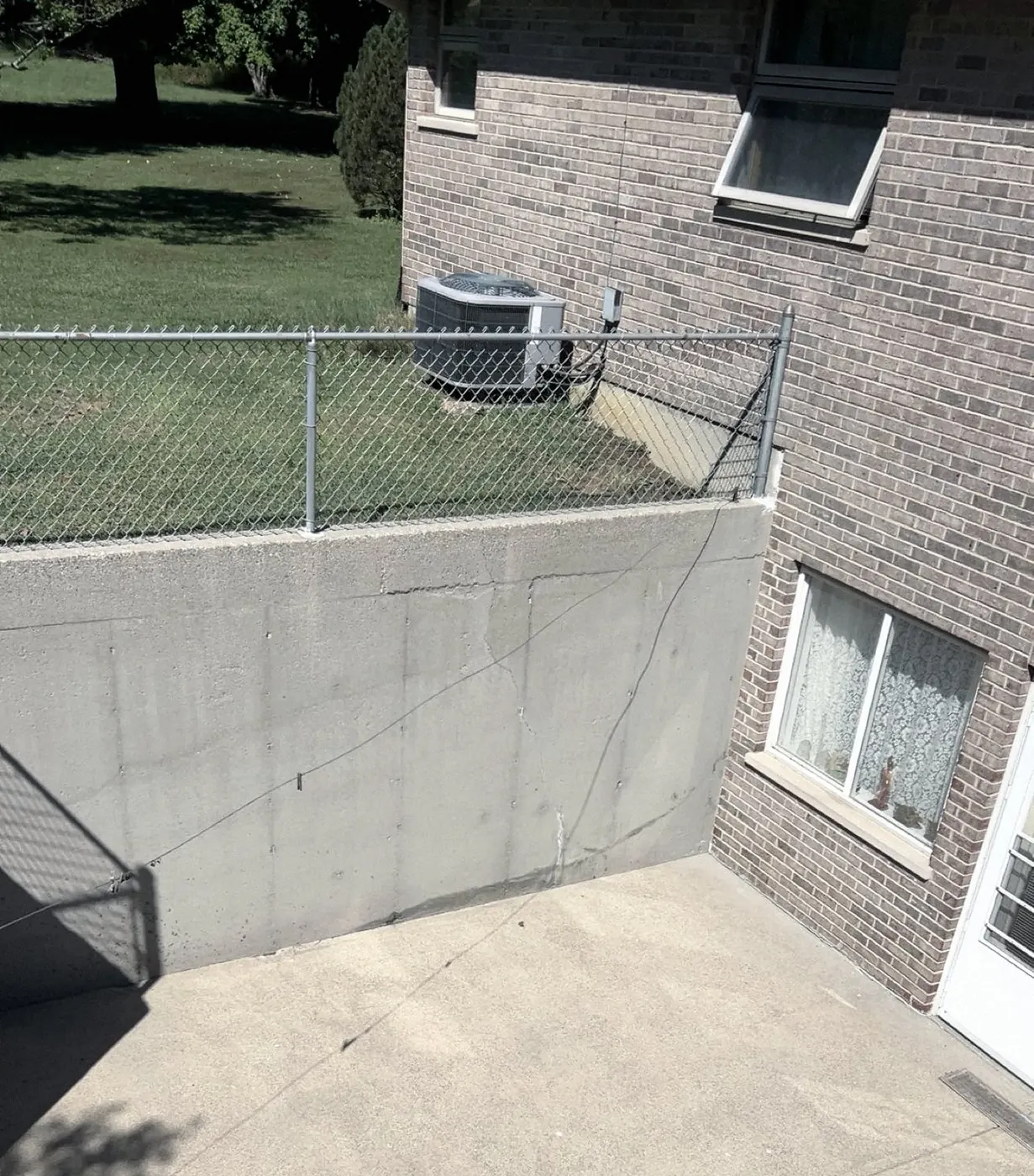 Light concrete patio and retaining wall with a chain-link fence, an AC unit and a brick wall with windows, shot from above.
