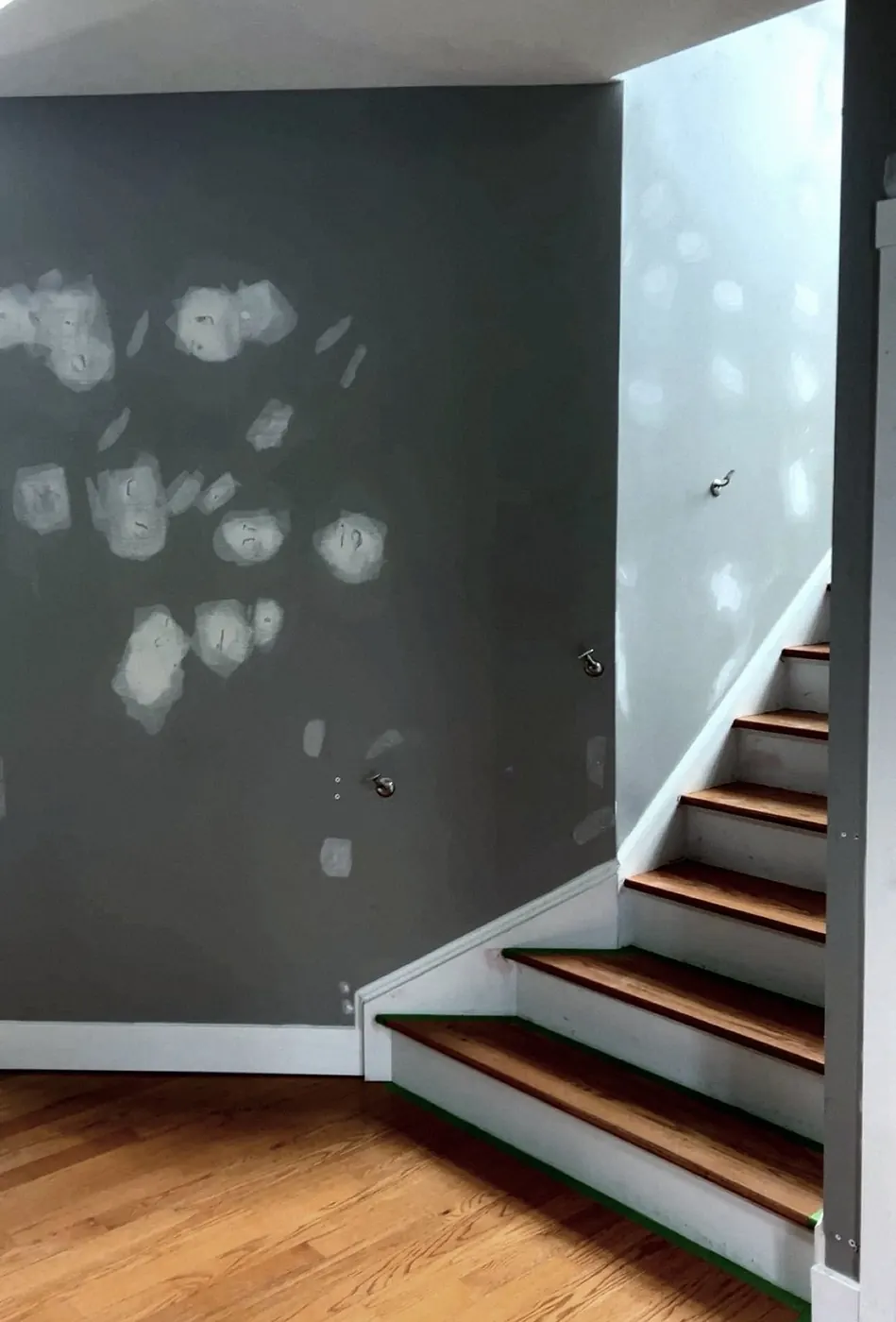 Gray stairwell wall dotted with white skim-coat patches, oak stair treads with green tape, a skylight edge at the top.