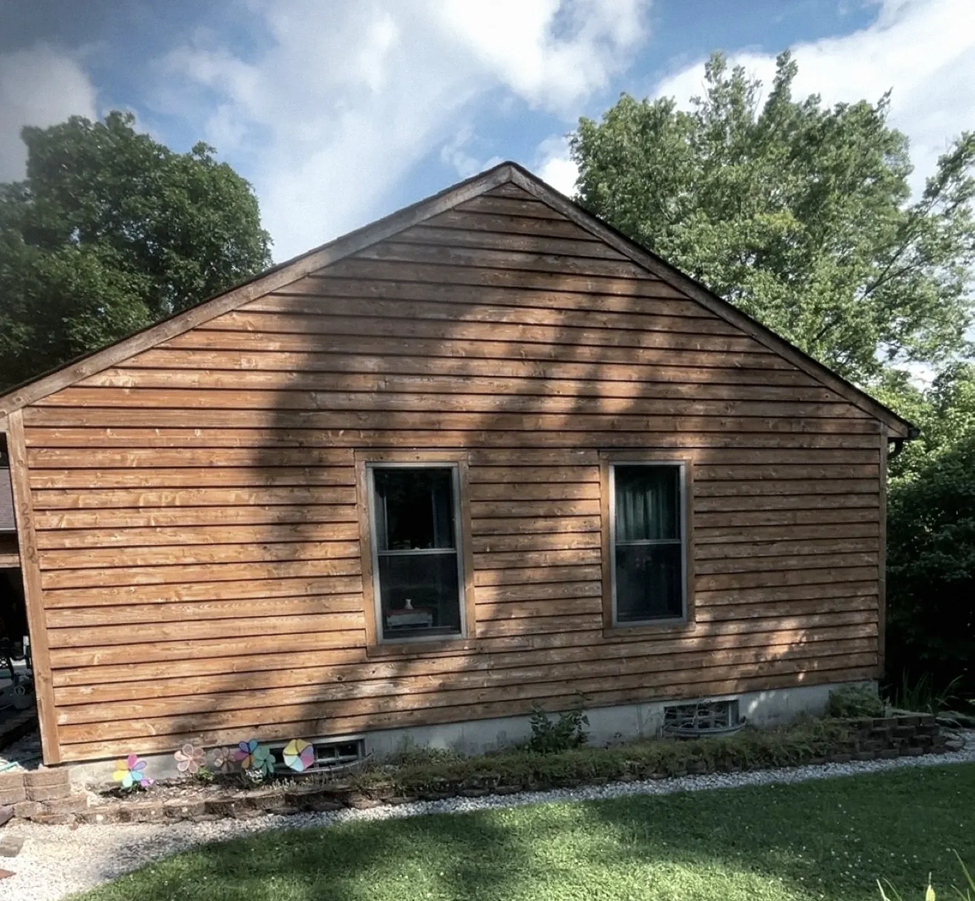 Side wall of a house in weathered orange-brown horizontal board siding with two windows, lawn in front, tree shadows across the boards.