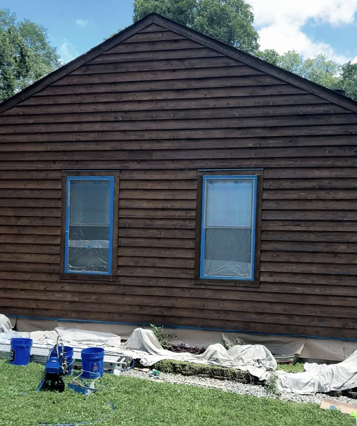 Siding stained dark brown, both windows masked in plastic with blue tape, an airless sprayer, buckets and drop cloths on the lawn.